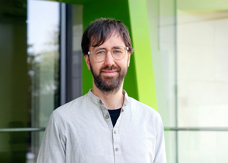 Prof. Dr. Mario Wenzel, neuer Professor für Gesundheitspsychologie an der Bergischen Universität Wuppertal, blickt lächelnd in die Kamera. Er trägt ein helles, kragenloses Hemd über einem dunklen T-Shirt und eine Brille. Das Porträt zeigt ihn vor einer modernen Glasfassade mit markanten grünen Architekturelementen auf dem Campus; der Hintergrund ist leicht unscharf