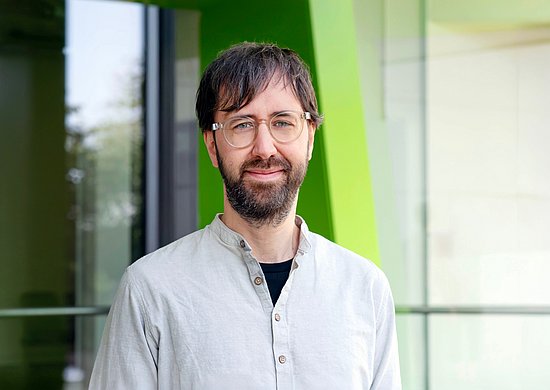 Prof. Dr. Mario Wenzel, neuer Professor für Gesundheitspsychologie an der Bergischen Universität Wuppertal, blickt lächelnd in die Kamera. Er trägt ein helles, kragenloses Hemd über einem dunklen T-Shirt und eine Brille. Das Porträt zeigt ihn vor einer modernen Glasfassade mit markanten grünen Architekturelementen auf dem Campus; der Hintergrund ist leicht unscharf