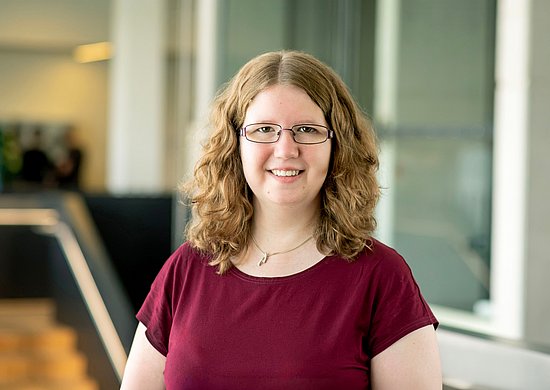 Porträtaufnahme von Dr. Marloes Venema. Junge Frau, hellbraunes, lockiges schulterlanges Haar, Brille, blickt lächelnd in die Kamera, trägt ein dunkelrotes Shirt.