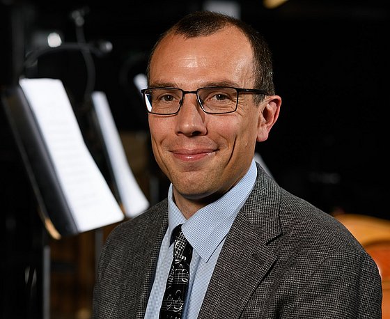 Das Porträt zeigt Georg Eckert. Der Wissenschaftler sitzt vor dunklem, leicht verschwommenem Hintergrund. Das Porträt zeigt Kopf und Oberkörper. Georg Eckert lächelt, trägt kurzes, dunkles Haar, eine schwarze Brille mit eckigen Gläsern, einen dunkelgraumelierten Anzug mit hellblauem Hemd und einer dunklen Krawatte mit hellen Mustern.