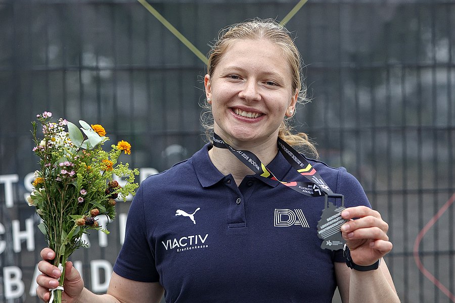 Das Bild zeigt Speerwerferin und BUW-Studentin Victoria Krause mit der gewonnenen Silbermedaille, die ihr um den Hals hängt und die sie mit der linken Hand festhält und in die Kamera zeigt. Die Medaille hat die Form von Deutschland und die Aufschrift "Deutsche Leichtathletik Meisterschaften". In der rechten Hand hält sie einen kleinen Blumenstrauß. Sie lacht in die Kamera.