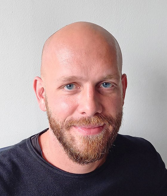 Mann mit Glatze und rot-braunem Vollbart. Er trägt ein schwarzes Shirt.