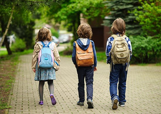 Drei Kinder mit Rucksack, zu sehen von hinten, gehen auf einem Weg mit Kopfsteinpflaster, im Hintergrund Bäume und Rasenfläche, am Ende des Weges unscharf angedeute Straßenkreuzung mit Autos.