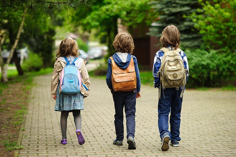 Drei Kinder mit Rucksack, zu sehen von hinten, gehen auf einem Weg mit Kopfsteinpflaster, im Hintergrund Bäume und Rasenfläche, am Ende des Weges unscharf angedeute Straßenkreuzung mit Autos.