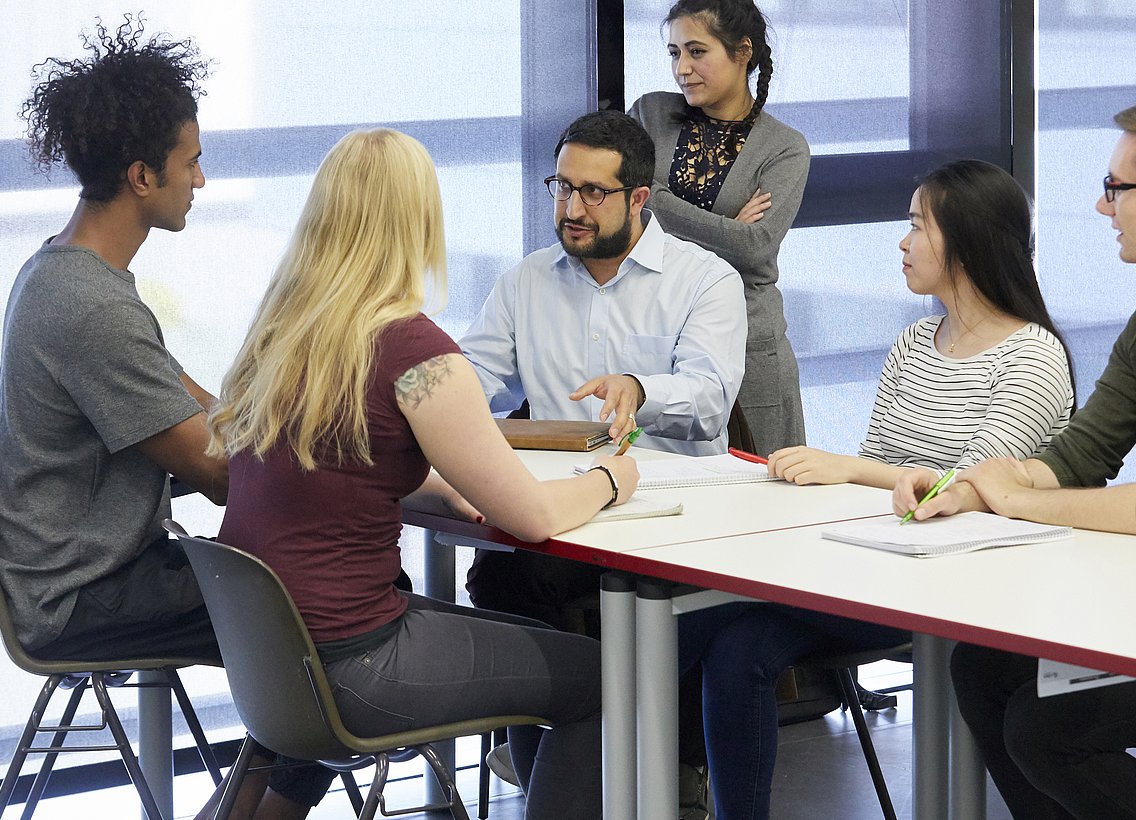Eine Gruppe von Studierenden sitzt um einen Tisch herum und ist in eine Gruppenaufgabe vertieft. Eine Person spricht, die anderen hören ihr aufmerksam zu.