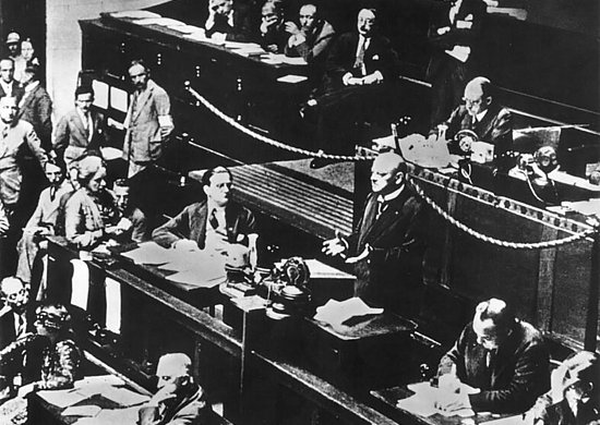 Historische Schwarz-Weiß-Aufnahme einer Sitzung des Völkerbunds. Mehrere Delegierte sitzen an Tischen im Sitzungssaal, während ein Redner im Vordergrund am Rednerpult steht.