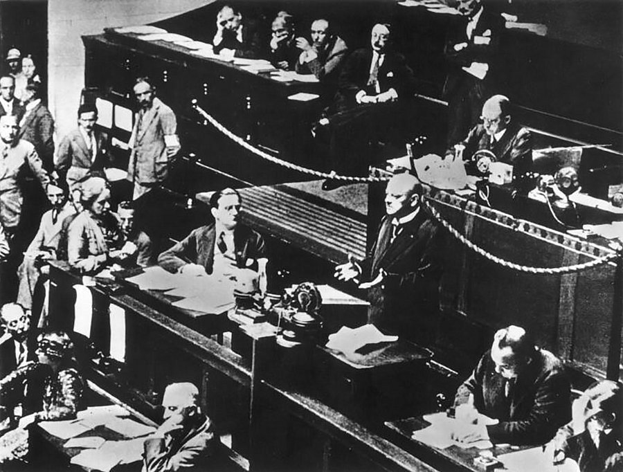 Historische Schwarz-Weiß-Aufnahme einer Sitzung des Völkerbunds. Mehrere Delegierte sitzen an Tischen im Sitzungssaal, während ein Redner im Vordergrund am Rednerpult steht.